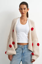 A woman modeling a cream-colored chunky knit cardigan with red pom-pom accents, worn over a white crop top and light blue jeans against a plain white background.