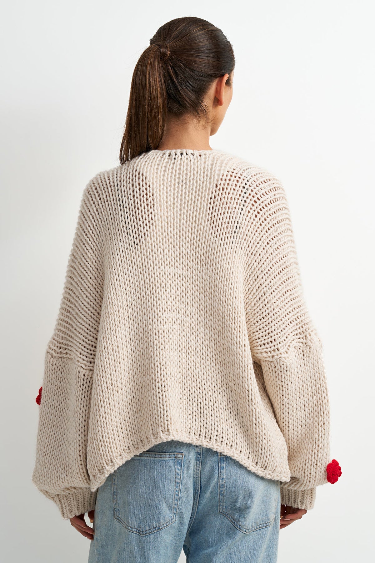 A woman viewed from behind wearing a chunky knit cream sweater with red pom-pom details on the sleeves, paired with light blue jeans against a plain white background.