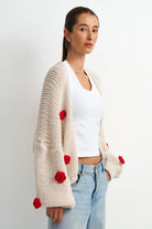 A woman wearing a chunky cream knit cardigan with red flower pom-pom details, styled over a white tank top and light blue jeans against a plain white background.