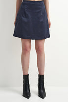 A navy blue A-line mini skirt with front pleats, shown on a model from the waist down wearing black ankle boots against a plain white studio background.