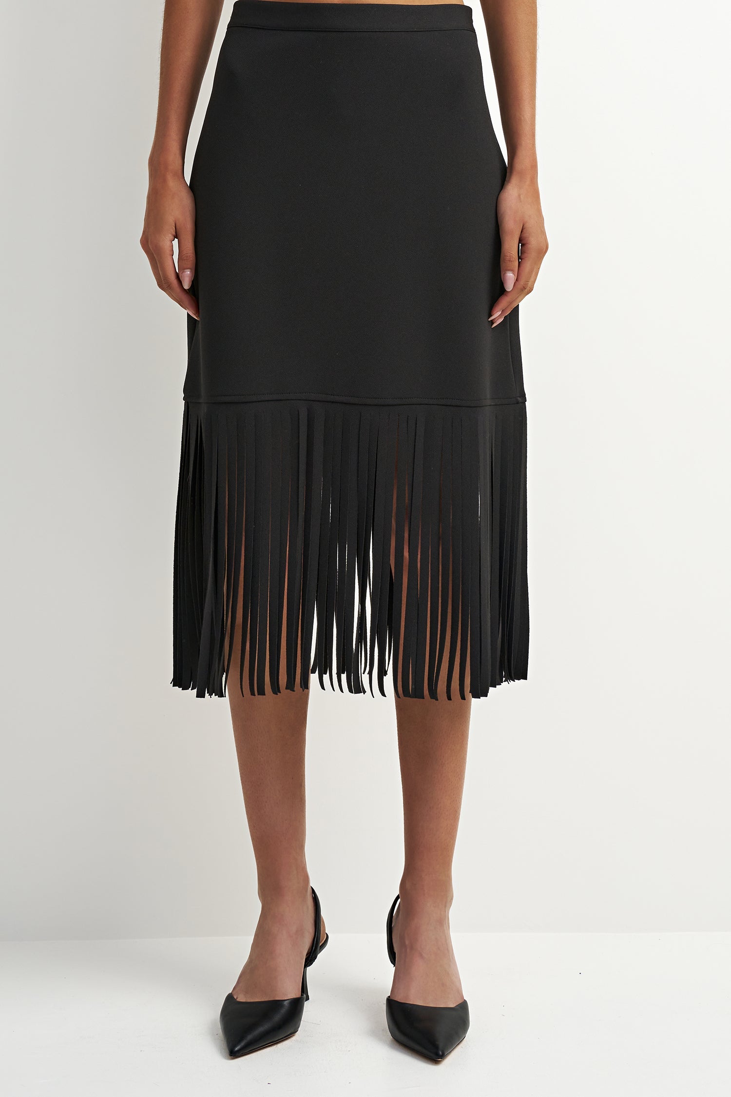 A black midi skirt with a tiered design and long fringe along the bottom hem, shown on a model wearing black pointed-toe heels against a plain white studio background.