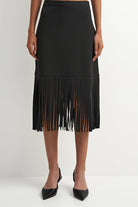A black midi skirt with a tiered design and long fringe along the bottom hem, shown on a model wearing black pointed-toe heels against a plain white studio background.