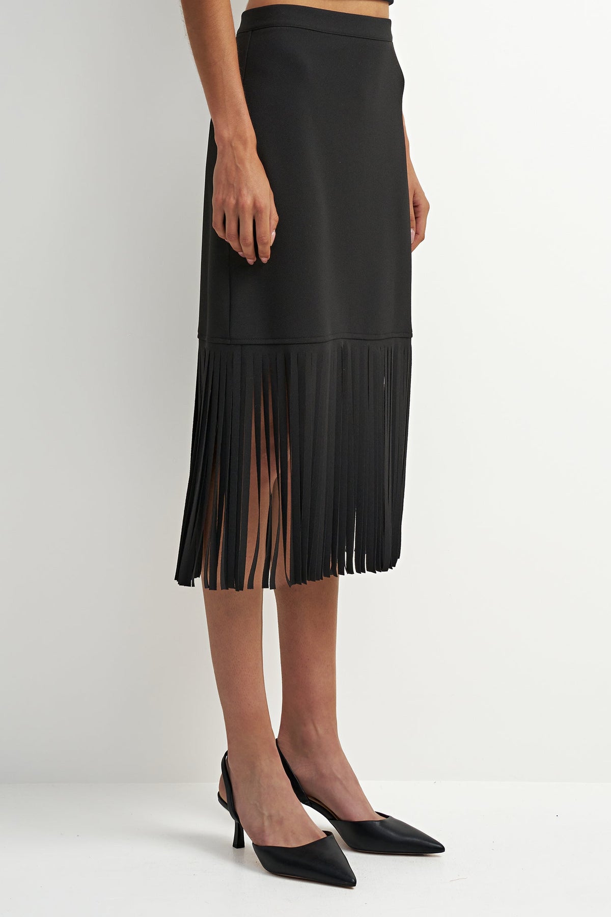 A black midi skirt with a fringe hem and side slit, shown on a model wearing black pointed-toe heels against a plain light gray studio background.