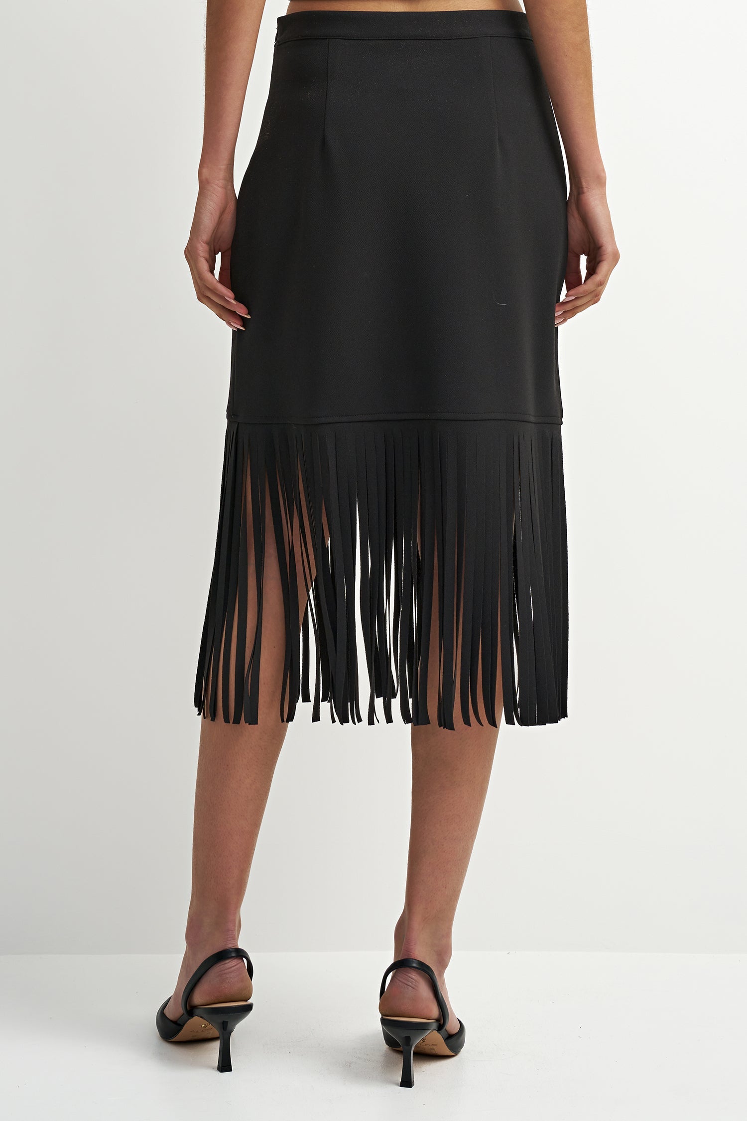 A black midi skirt with a long fringe hem, shown from the back on a model wearing black strappy heels against a plain white studio background.
