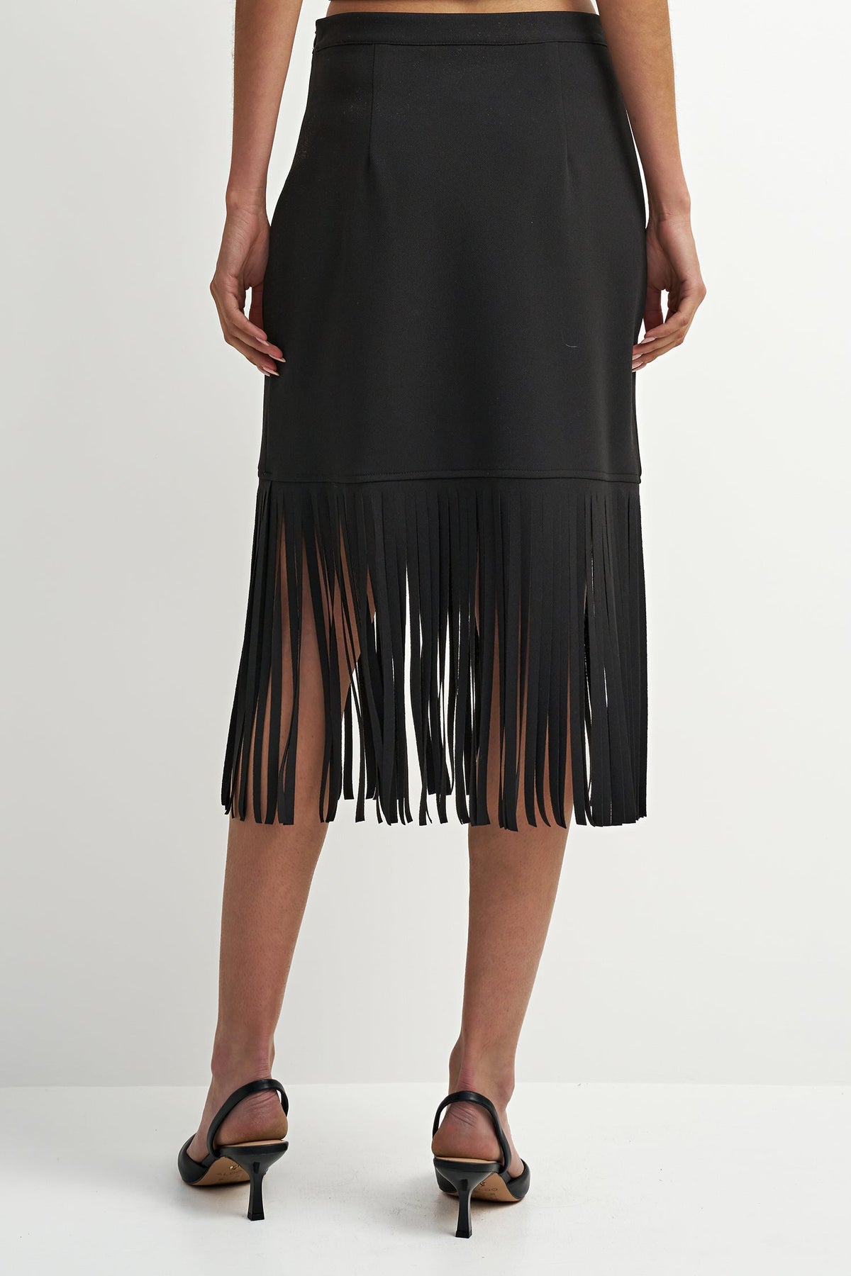 A black midi skirt with a long fringe hem, shown from the back on a model wearing black strappy heels against a plain white studio background.
