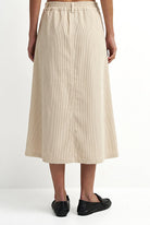 A model from the waist down wearing a light beige linen midi skirt with an elastic waistband and black flat loafers, set against a plain white studio background.