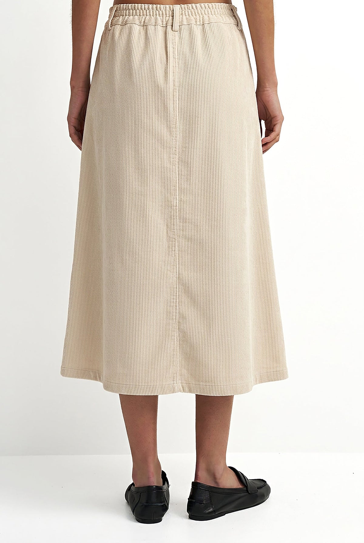 A model from the waist down wearing a light beige linen midi skirt with an elastic waistband and black flat loafers, set against a plain white studio background.