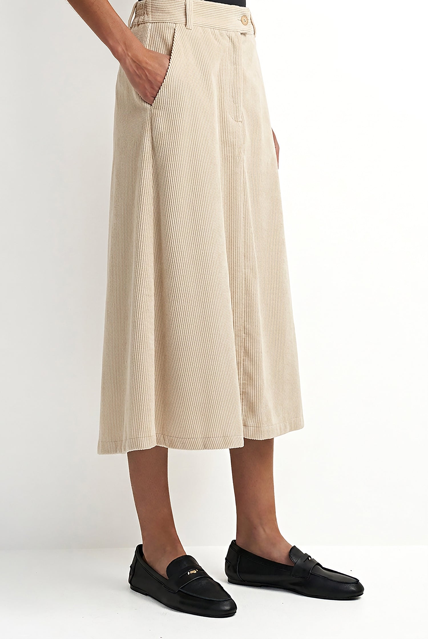 A model wearing a high-waisted, wide-leg midi skirt in beige corduroy, paired with black leather loafers, shown from the waist down against a plain white studio background.