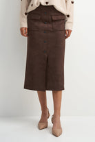 A model wearing a dark brown midi skirt made of a soft suede-like material, paired with a cream knit cardigan and beige pointed-toe heels, set against a plain light gray studio background.