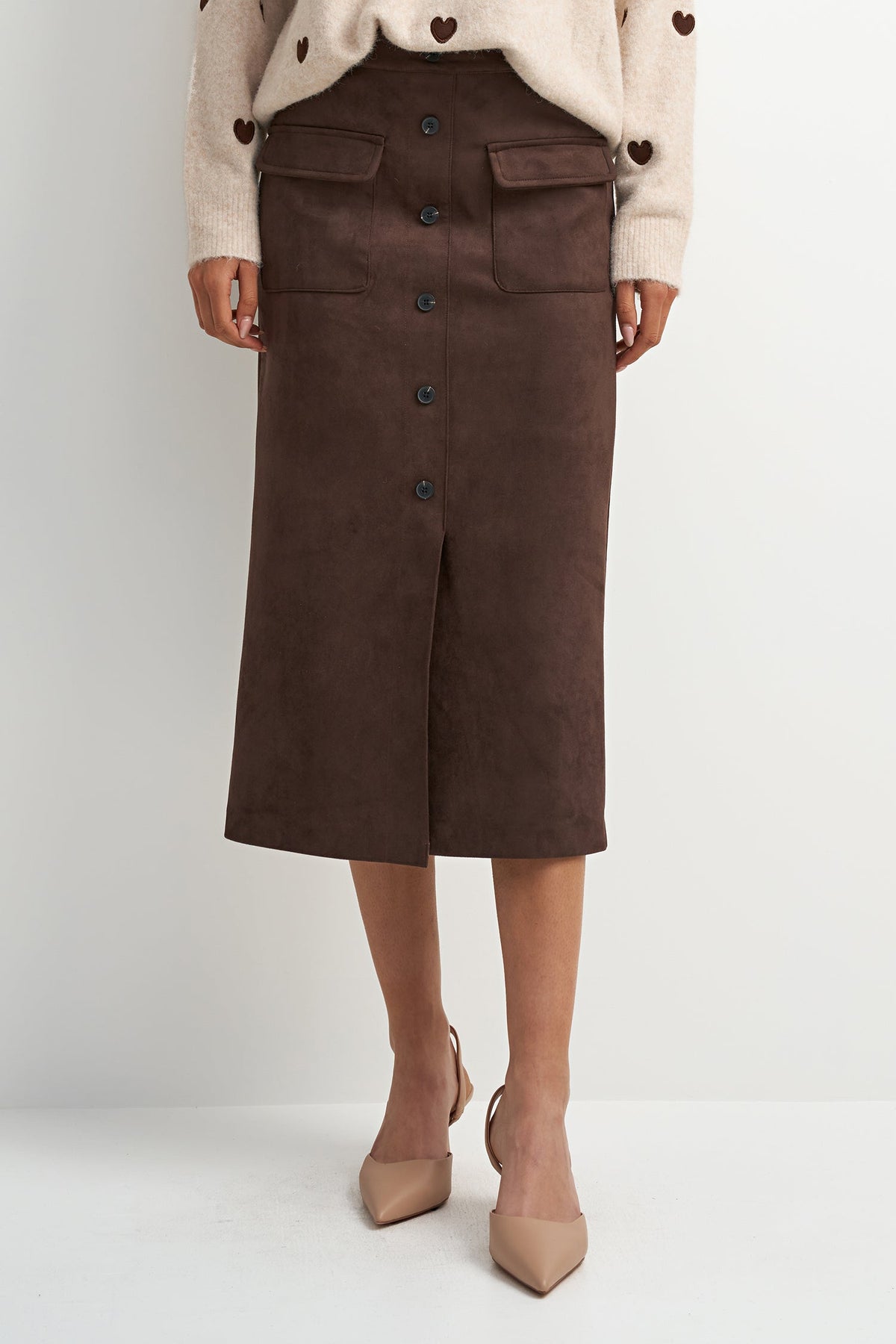 A model wearing a dark brown midi skirt made of a soft suede-like material, paired with a cream knit cardigan and beige pointed-toe heels, set against a plain light gray studio background.