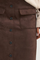 Close-up of a brown suede-like button-front garment with flap chest pockets and a cream ribbed sleeve or sweater visible at the top and right edge.
