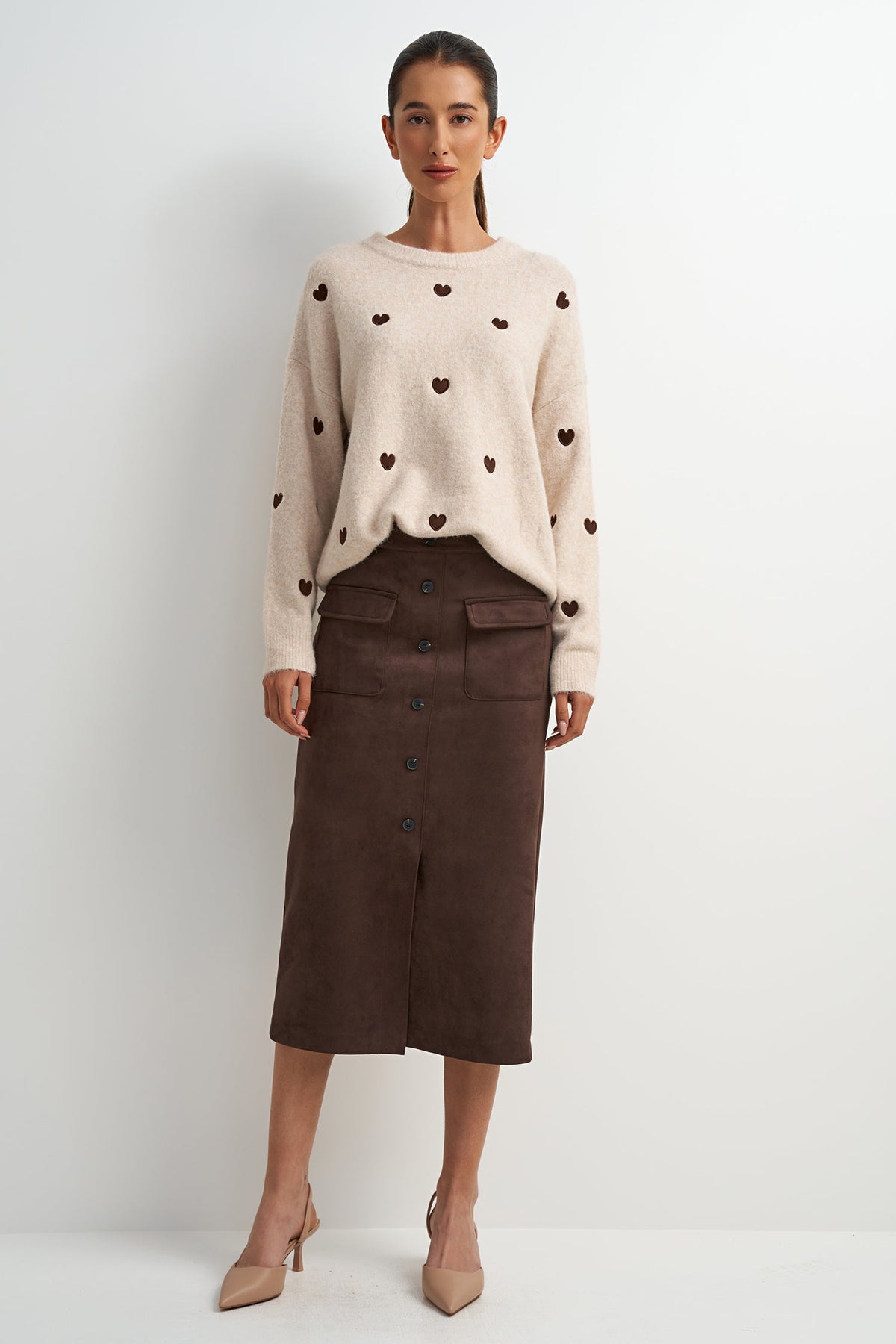 A model stands against a light gray studio background wearing a cream knit sweater with small dark heart motifs and a dark brown midi skirt.