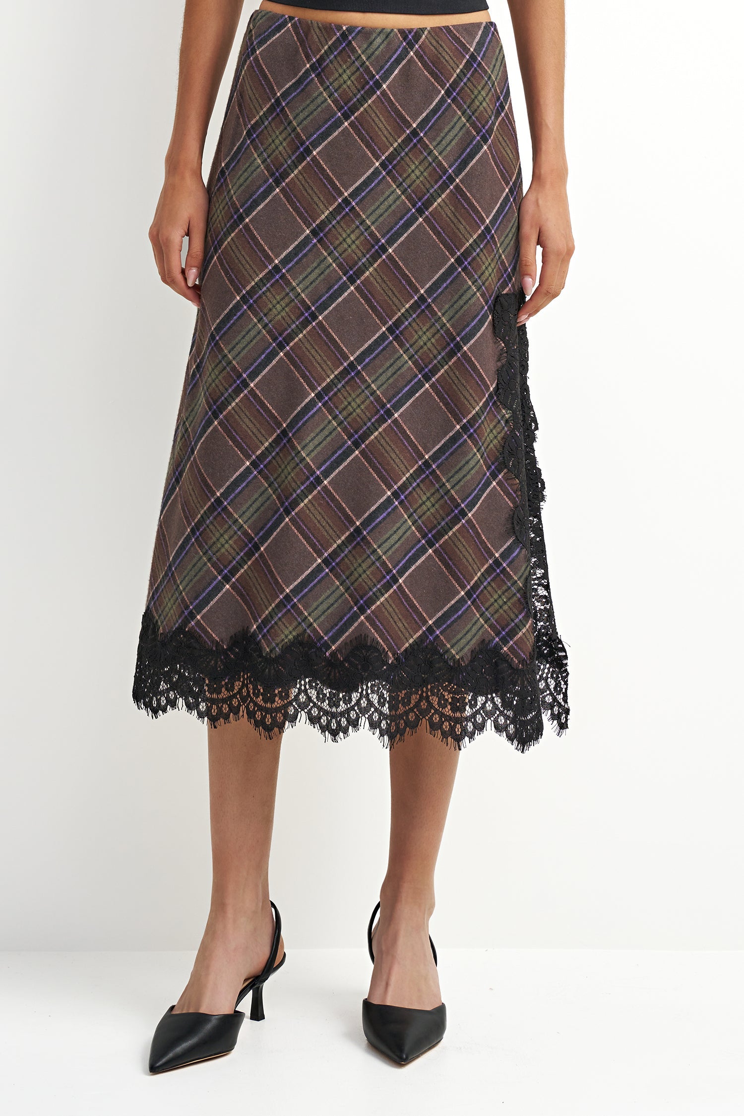 A woman models a brown plaid midi skirt with black lace trim at the hem and side, paired with black pointed-toe heels against a plain white studio background.
