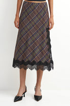 A woman models a brown plaid midi skirt with black lace trim at the hem and side, paired with black pointed-toe heels against a plain white studio background.
