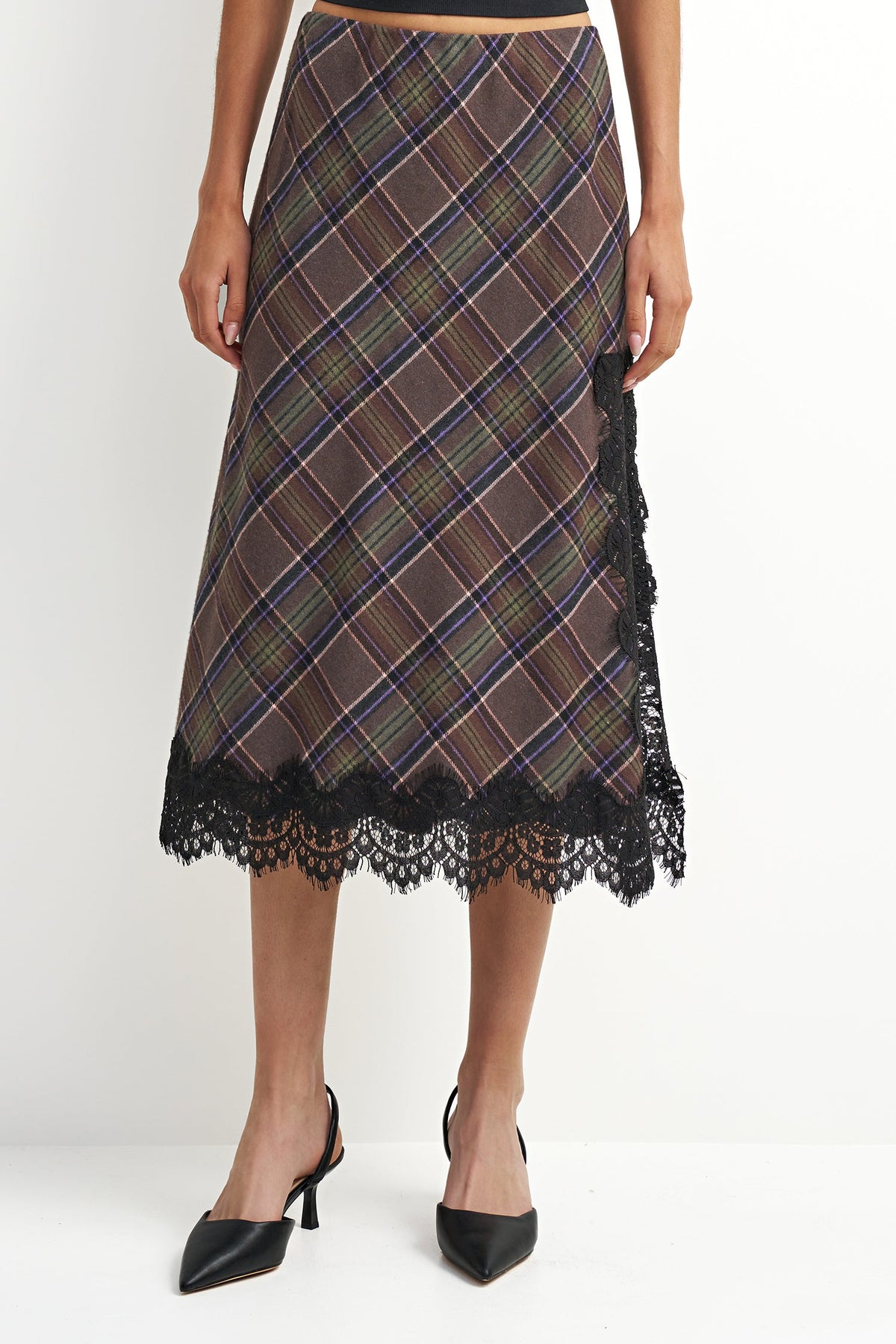 A woman models a brown plaid midi skirt with black lace trim at the hem and side, paired with black pointed-toe heels against a plain white studio background.