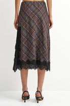 A woman's midi skirt with a brown, green, and blue plaid pattern and black lace trim, shown from the back on a model wearing black slingback heels against a white studio background.