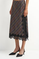 A model wearing a brown and black plaid midi skirt with black lace trim, paired with black pointed-toe heels, shown from the waist down against a plain white studio background.