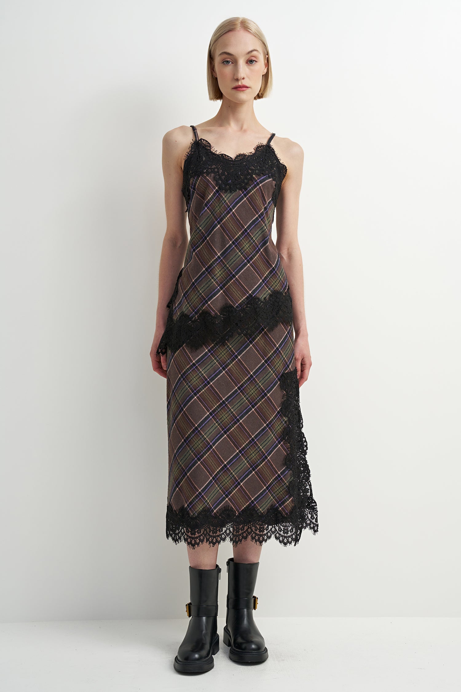 A model wearing a sleeveless midi dress with a brown and black plaid pattern, black lace trim, and black ankle boots, standing against a plain white studio background.