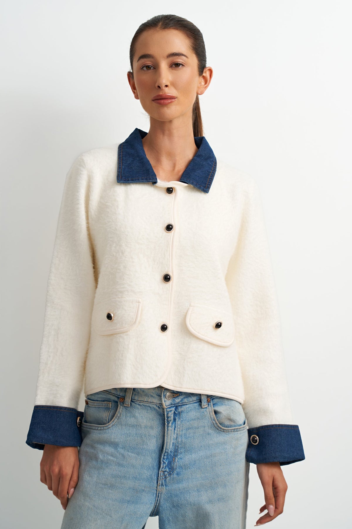 A woman modeling a cropped cream-colored textured jacket with contrasting dark blue collar, cuffs, and buttons, paired with light-wash blue jeans against a plain light gray background.
