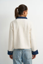 A woman viewed from the back wearing a cream-colored fuzzy cropped cardigan with dark blue collar and cuff accents, paired with light blue jeans, against a plain light gray background.