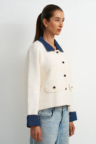 A woman wearing a cropped cream-colored tweed jacket with contrasting navy blue collar, cuffs, and buttons, paired with light-wash blue jeans, shown from a side profile against a light gray studio background.