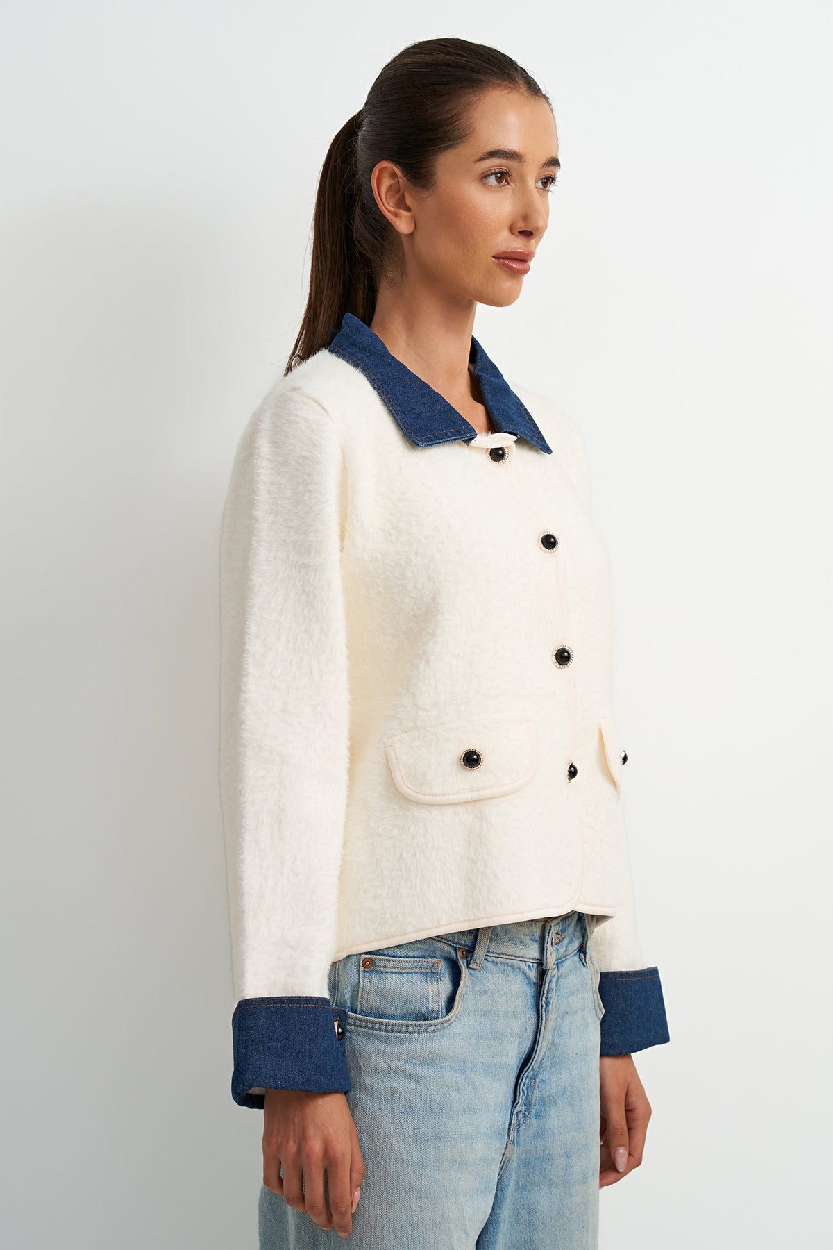 A woman wearing a cropped cream-colored tweed jacket with contrasting navy blue collar, cuffs, and buttons, paired with light-wash blue jeans, shown from a side profile against a light gray studio background.