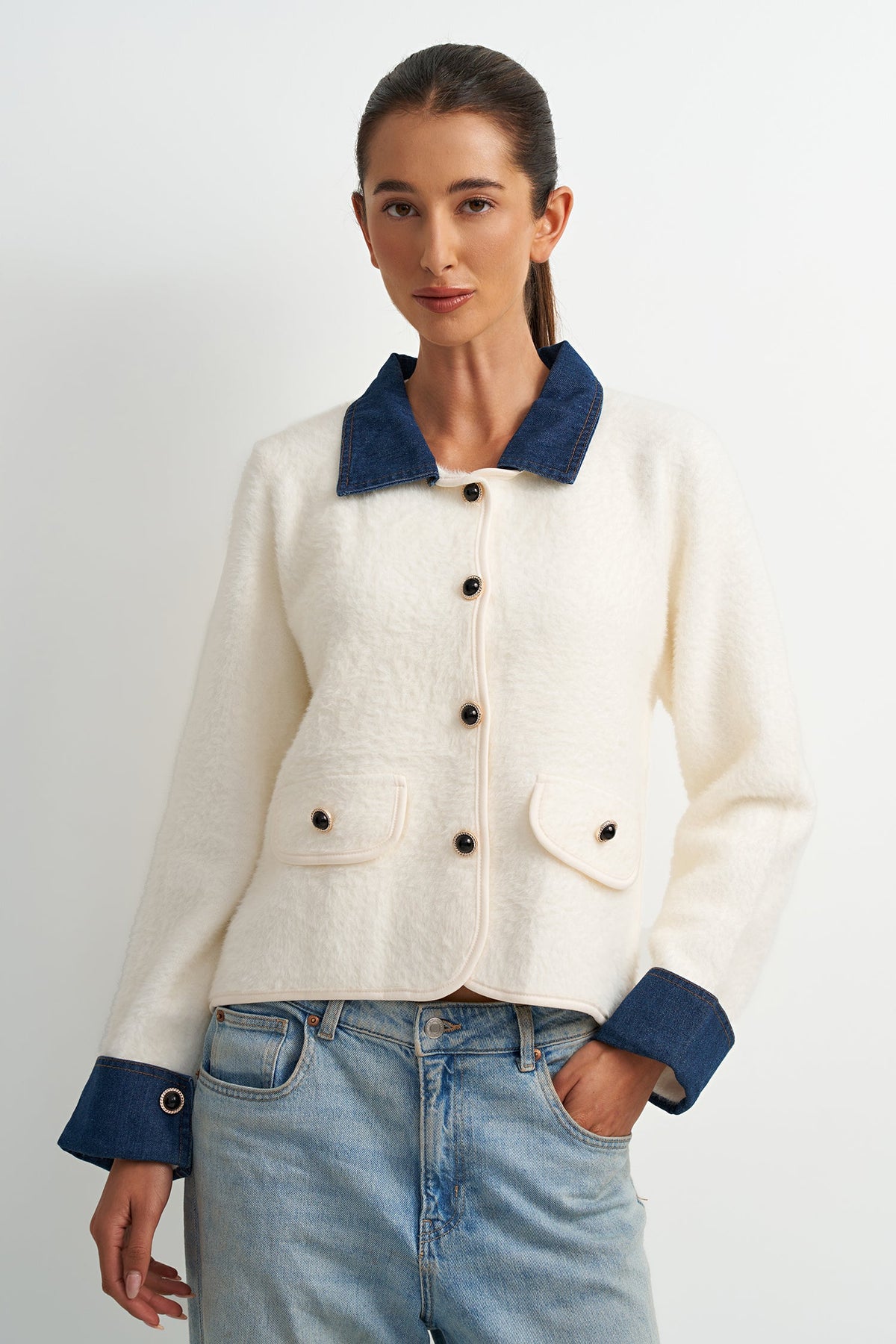 A woman modeling a cropped cream-colored textured jacket with contrasting dark blue collar, cuffs, and button details, paired with light-wash blue jeans against a plain light gray background.