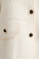 Close-up of a cream-colored textured faux fur jacket featuring a pocket with a black button and gold-tone trim.