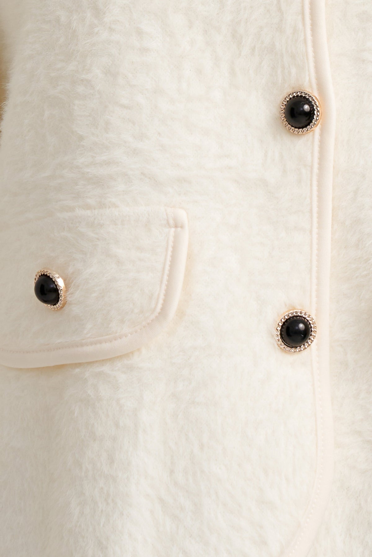 Close-up of a cream-colored textured faux fur jacket featuring a pocket with a black button and gold-tone trim.