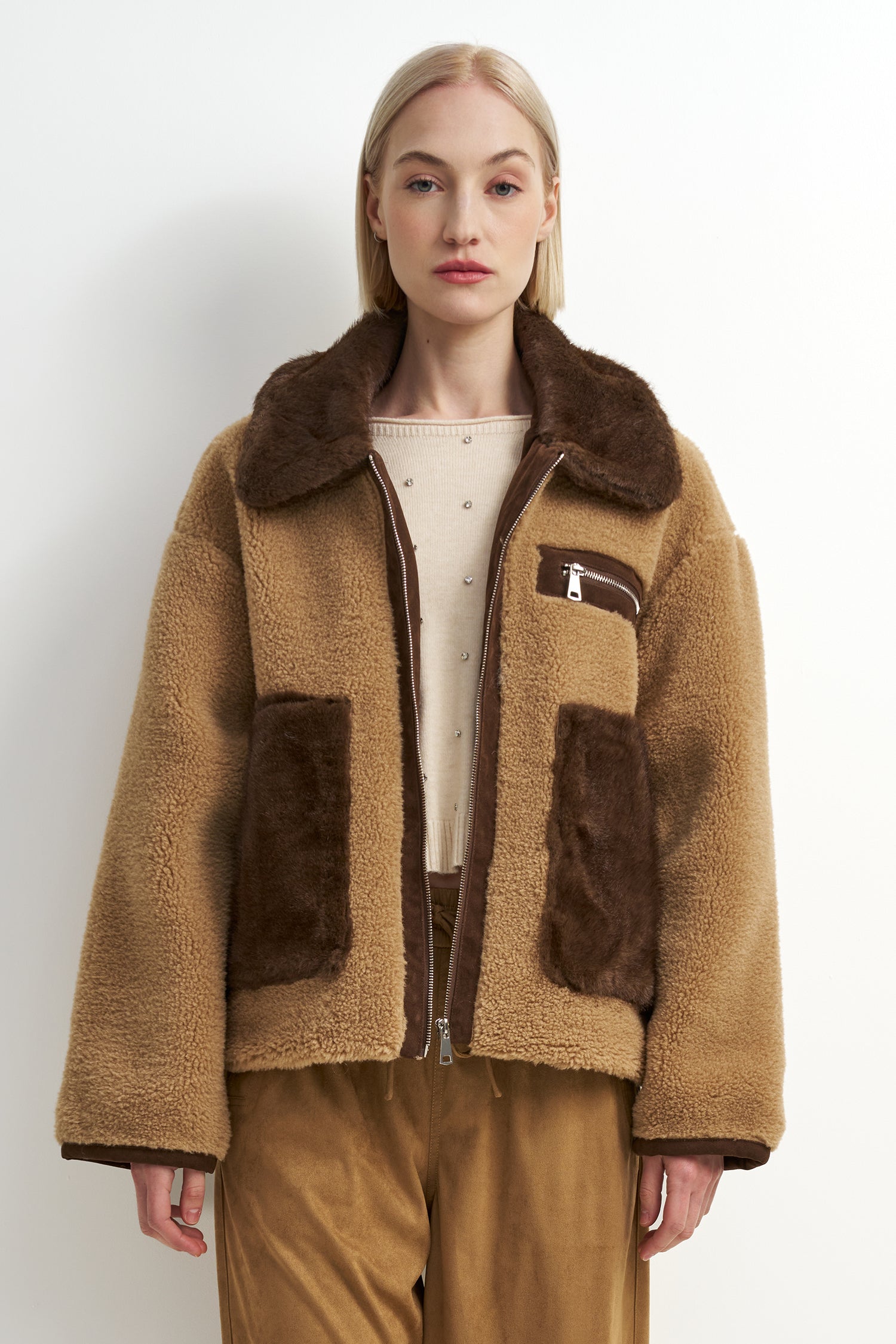 A model wearing a tan and brown shearling-style jacket with a large brown collar, brown patch pockets, and a front zipper, paired with tan pants against a plain light background.