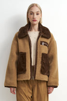 A model wearing a tan and brown shearling-style jacket with a large brown collar, brown patch pockets, and a front zipper, paired with tan pants against a plain light background.