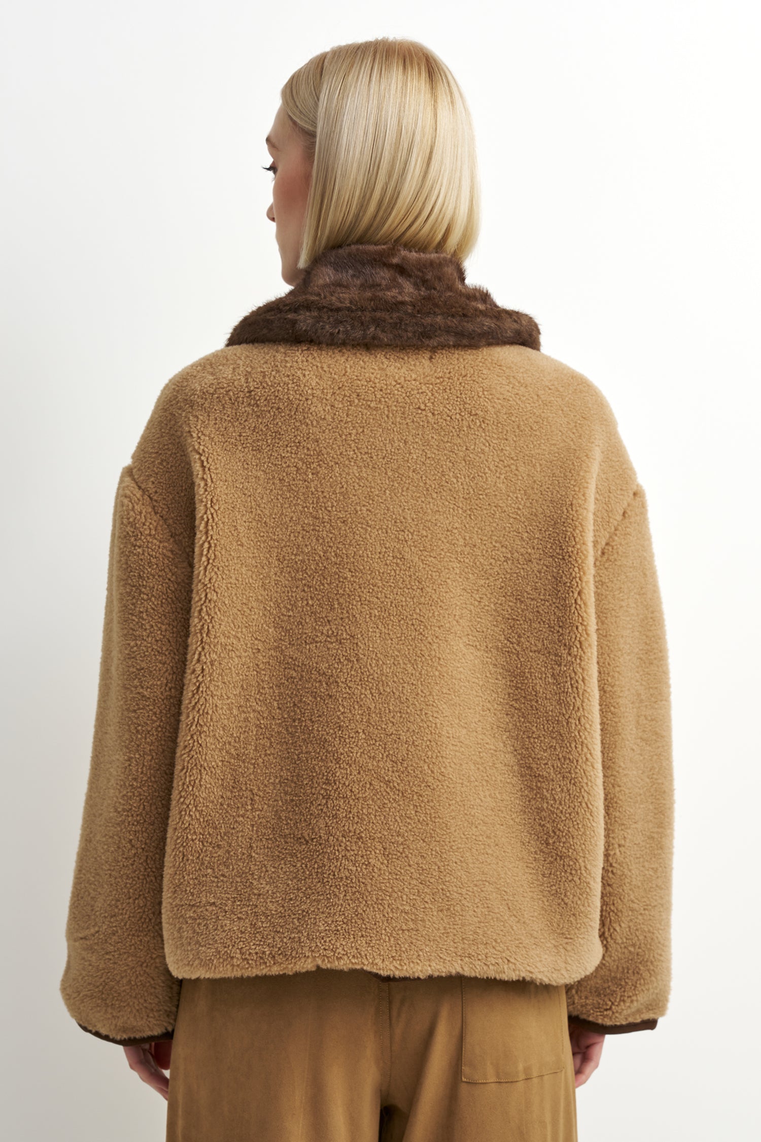 A model shown from the back wearing a camel brown sherpa fleece jacket with a contrasting dark brown faux fur collar, paired with matching brown trousers, against a plain white background.