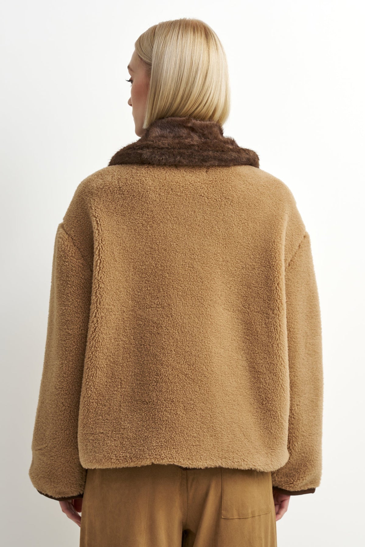 A model shown from the back wearing a camel brown sherpa fleece jacket with a contrasting dark brown faux fur collar, paired with matching brown trousers, against a plain white background.