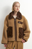 A model wearing a tan sherpa fleece jacket with dark brown faux-fur trim, a dark brown zipper, and two large dark brown patch pockets, paired with brown pants against a plain white studio background.