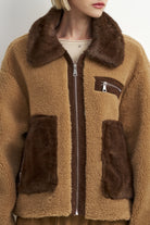 A cropped view of a person wearing a brown sherpa fleece zip-up jacket with dark brown faux-fur trim on the collar and front pockets, over a light beige top.