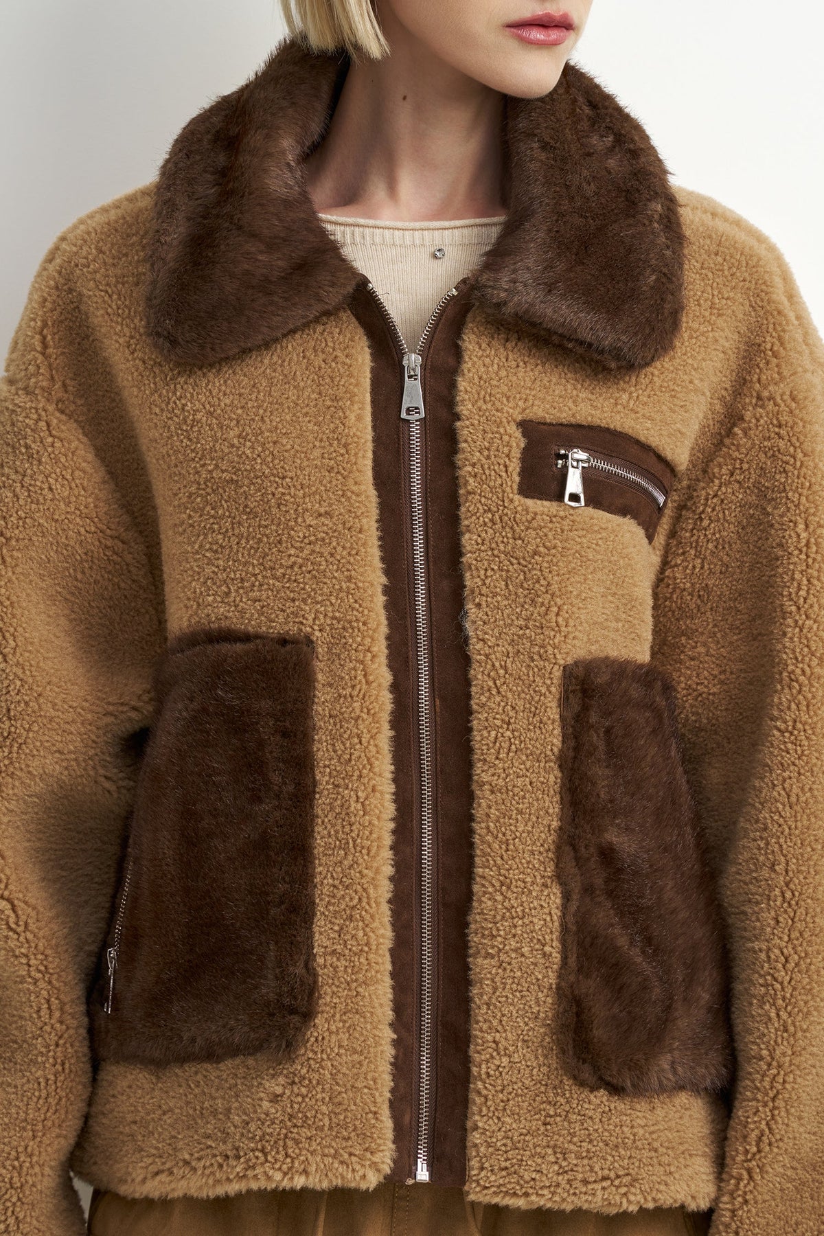 A cropped view of a person wearing a brown sherpa fleece zip-up jacket with dark brown faux-fur trim on the collar and front pockets, over a light beige top.