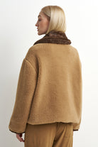 A woman shown from the back wearing a camel-colored teddy coat with a dark brown faux-fur collar, paired with matching brown trousers, against a plain white studio background.