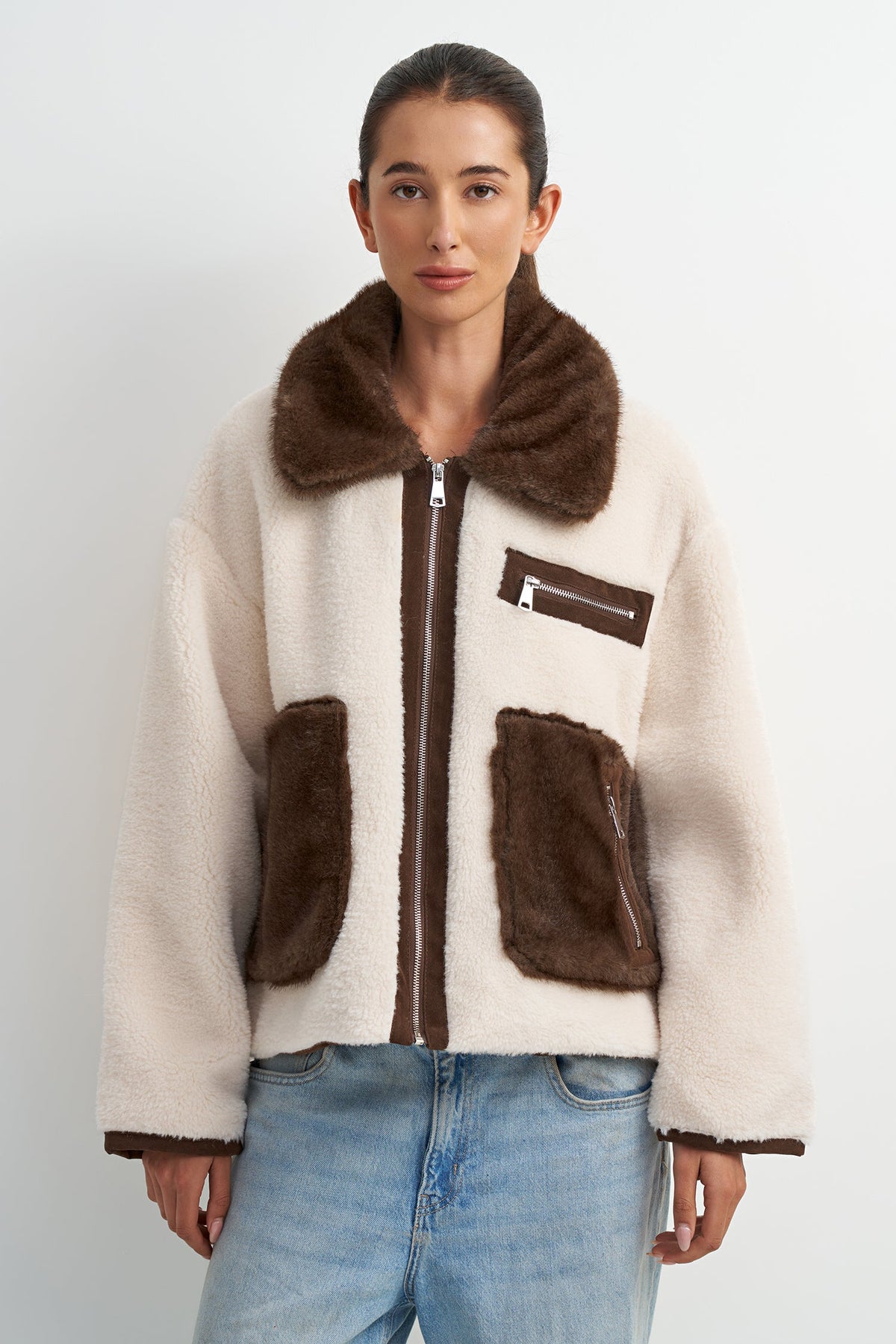 A woman wearing a cream-colored shearling-style jacket with dark brown faux-fur trim, a brown collar, and two large brown front pockets, paired with light blue jeans, against a light gray studio background.