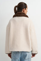 A woman shown from the back wearing a cream-colored fuzzy teddy jacket with a brown faux-fur collar, paired with light blue jeans, against a plain white studio background.
