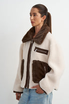 A woman wearing a cream-colored shearling-style jacket with dark brown faux-fur trim, a brown collar, and brown zippered pockets, paired with light blue jeans, against a plain light gray background.