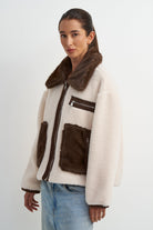 A woman wearing a cream-colored faux fur jacket with dark brown fur trim, pockets, and zipper details, paired with light blue jeans, against a plain light gray background.