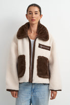 A woman modeling a cream-colored sherpa jacket with dark brown faux-fur trim, a brown collar, and two large brown front pockets, paired with light blue jeans against a plain light gray background.