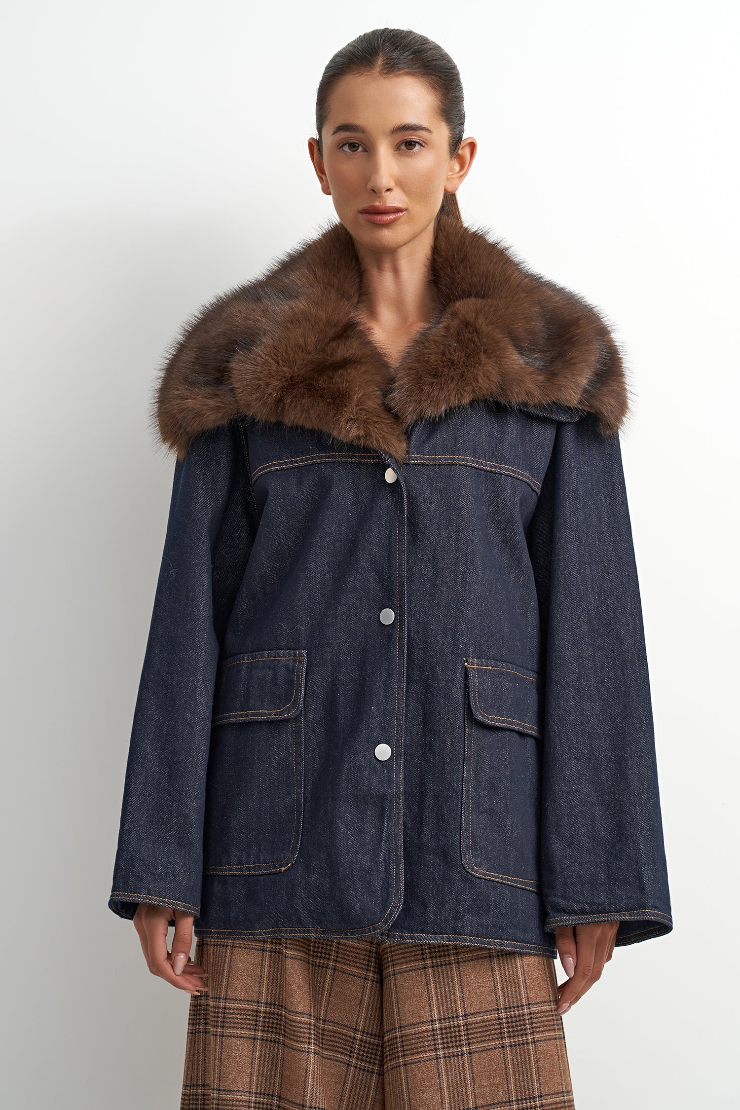 A model wearing a dark blue denim jacket with a large brown fur collar, paired with brown plaid trousers, against a plain light gray background.