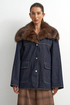 A model wearing a dark blue denim jacket with a large brown fur collar, paired with brown plaid trousers, against a plain light gray background.