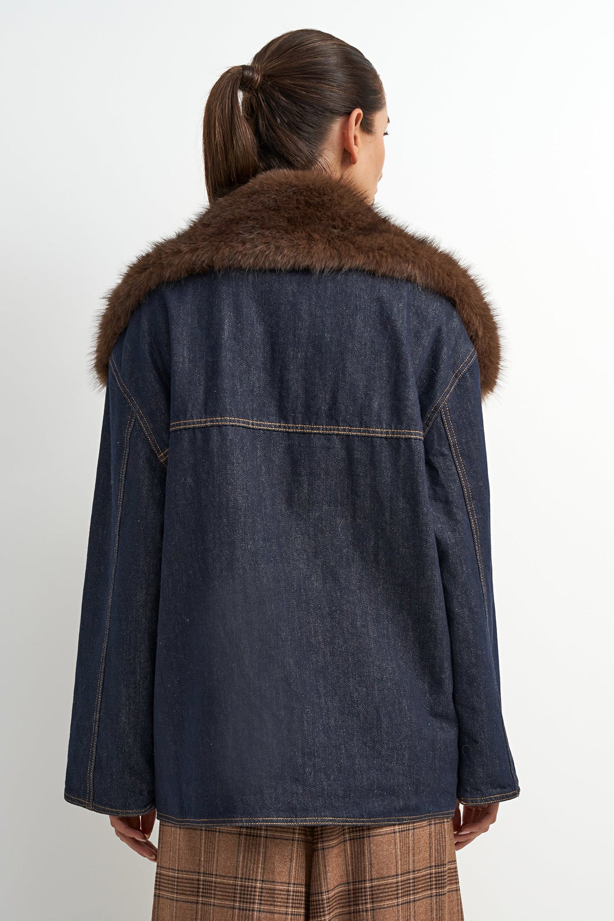 A woman shown from the back wearing a dark blue denim jacket with a brown faux-fur collar, paired with brown plaid pants, against a plain white background.