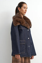 A woman wearing a dark blue denim jacket with a large brown fur collar, shown in profile against a plain light gray background.