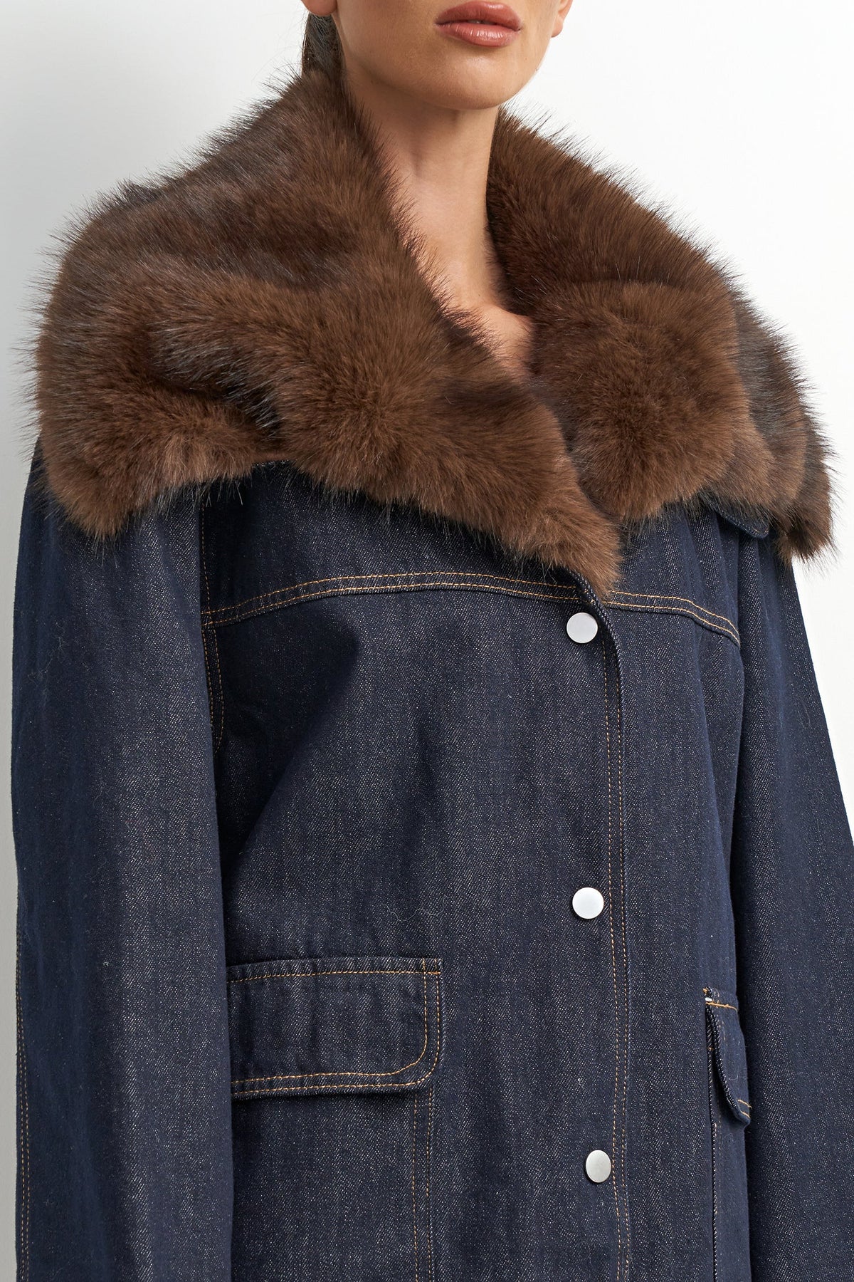 A close-up of a model wearing a dark blue denim jacket with a large, fluffy brown fur collar. The jacket features silver buttons down the front and patch pockets.