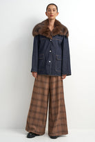A model stands against a plain light gray studio background wearing a dark blue denim jacket with a large brown fur collar, paired with brown plaid wide-leg trousers and black shoes.