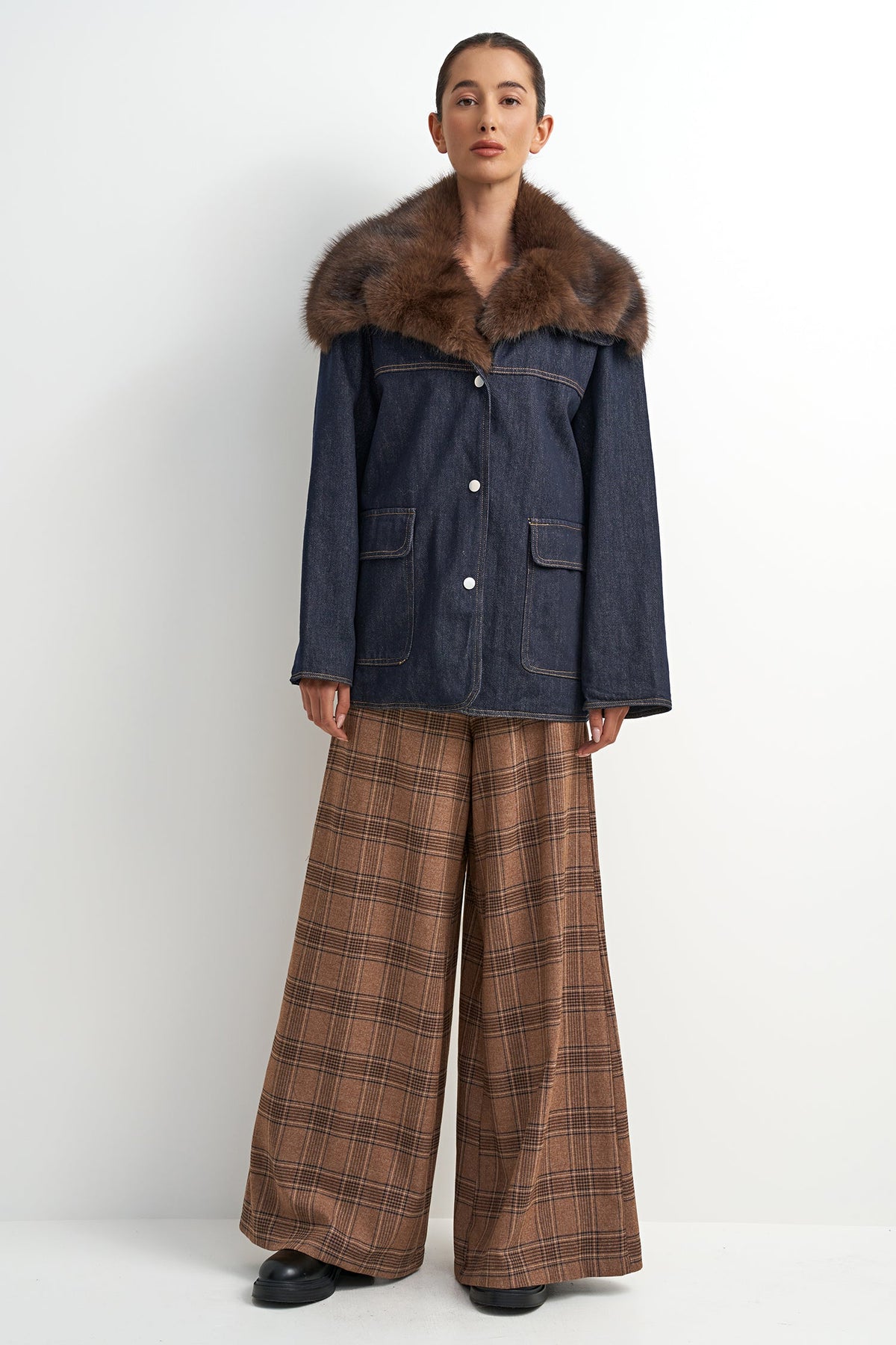 A model stands against a plain light gray studio background wearing a dark blue denim jacket with a large brown fur collar, paired with brown plaid wide-leg trousers and black shoes.
