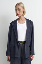 A model wearing a navy blue pinstripe blazer and matching trousers over a white crewneck t-shirt, styled with a black belt with a gold buckle, against a plain light gray background.